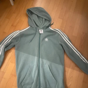 Grön huvtröja från Adidas - Säljer en grön huvtröja från Adidas med klassiska vita ränder längs ärmarna. Tröjan har dragkedja och en broderad logga på bröstet. Perfekt för en avslappnad stil.
