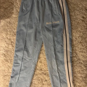 Ljusa blå byxor från Palm Angels - Snygga ljusblå byxor från Palm Angels med vita och svarta ränder längs sidorna. Byxorna har elastisk midja och sidofickor, perfekt för en avslappnad stil. Märkesloggan är tryckt på framsidan.