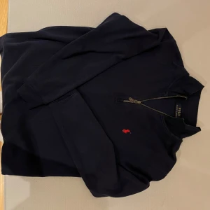 Svart tröja med dragkedja från Polo Ralph Lauren - Snygg svart tröja från Polo Ralph Lauren med en liten röd broderad logga på bröstet. Tröjan har en dragkedja vid halsen och långa ärmar, perfekt för en stilren look.