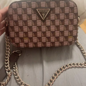 Brun axelväska från Guess - Snygg brun axelväska från Guess med ett geometriskt mönster och kedjedetaljer. Väskan har en dragkedja upptill och är perfekt för att bära dina nödvändigheter med stil.