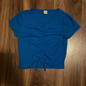 Blå topp  - Säljer en blå t-shirt/topp från Lindex i storlek 158/164 (12-14år) tröjan har en knuten rosett på framsidan. Den är även ribbad i tyget. 