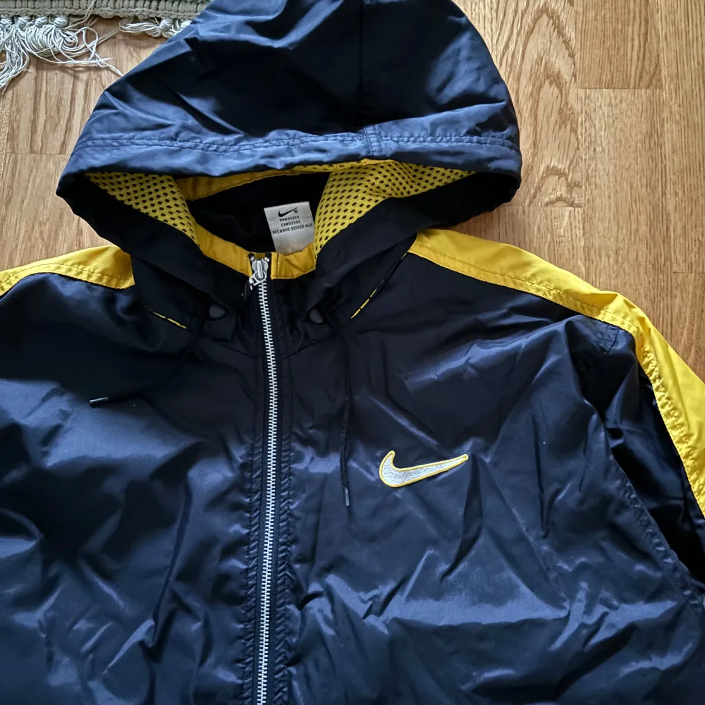 Snygg svart och gul vindjacka från Nike med huva och dragkedja. Jackan har långa ärmar och ett stort Nike-logotyp på ryggen. Perfekt för blåsiga dagar.. Takit.
