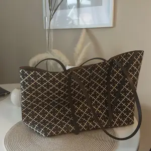 Snygg shoppingväska med ett stilrent rutigt mönster i brun och beige. Väskan har långa, smala bruna handtag och en rymlig insida, perfekt för shoppingturen. Den är både praktisk och trendig.