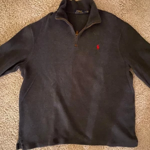 Grå tröja med dragkedja från Ralph Lauren - Säljer en stilren ljus svart tröja från Ralph Lauren med halv dragkedja och röd logga på bröstet. Perfekt för kyligare dagar med sin mjuka och bekväma passform.