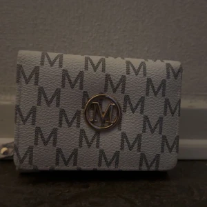 Vit och grå plånbok med M-mönster - Snygg plånbok i vitt och grått med ett stilrent M-mönster. Den har en praktisk dragkedja och en elegant guldfärgad logotyp på framsidan. Perfekt för att hålla ordning på kort och kontanter. Passar både till vardags och fest!