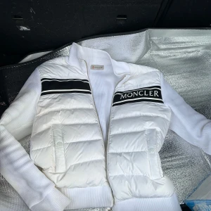 Vit cardigan jacka från Moncler - Säljer en snygg vit pufferjacka från Moncler i mycket bra skick. Jackan har en stilren design med svarta detaljer och Moncler-logga på bröstet. Perfekt för kyliga dagar med sin quiltade stil och dragkedja framtill. Passar både till vardags och festliga tillfällen.