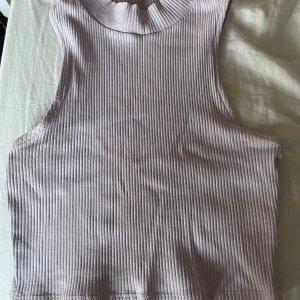 Ljuslila ribbad ärmlös topp - Säljer en ljuslila ribbad topp i ärmlös design. Perfekt för sommardagar eller som ett lager under en jacka. Toppen har en hög halsringning och är i bra skick, då den har visa defekter på sig som visas i bilden. Passar både till vardags och festliga tillfällen.
