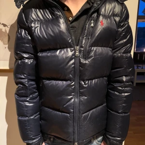 Svart pufferjacka med huva - Snygg svart pufferjacka från Polo Ralph Lauren med huva och dragkedja. Jackan har en broderad logga på bröstet och är perfekt för kyliga dagar. Den är långärmad och har en stilren design. Liten defekt där nere på jackan men inget stort.