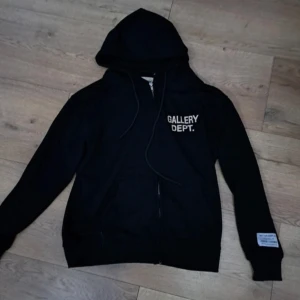 Svart hoodie från Gallery Dept. - Säljer en svart hoodie från Gallery Dept. med dragkedja och tryck på bröstet. Den har en justerbar huva och en unik etikett på ärmen. Perfekt för en avslappnad stil.