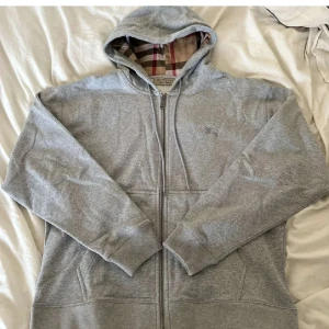Grå hoodie från Burberry - Säljer en stilren grå hoodie från Burberry med klassiskt rutigt mönster i huvan. Tröjan har dragkedja framtill och långa ärmar. Perfekt för en avslappnad och trendig look.