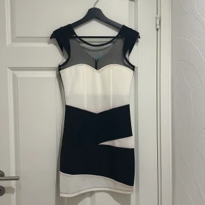 Svart och vit klänning med dragkedja - Snygg svart och vit klänning med geometriskt mönster och transparenta detaljer vid axlarna. Klänningen har en dragkedja baktill och är ärmlös. Perfekt för en stilren look.