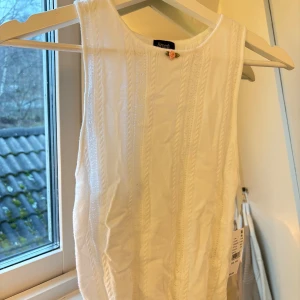 Vit ärmlös topp från Beverly and Beck - Säljer en söt vit ärmlös topp från Beverly and Beck. Den har en fin liten rosdetalj vid halsen och är i nytt skick. Toppen är gjord i ett mjukt material.