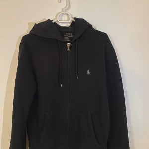 Svart hoodie från Polo Ralph Lauren - Säljer en stilren svart hoodie från Polo Ralph Lauren. Den har en dragkedja framtill och en klassisk logga på bröstet. Perfekt för både vardag och chill. Den är i bra skick och superbekväm att ha på sig. Passar perfekt till höst och vår!
