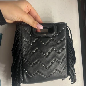 Svart handväska med fransar från Maje - Snygg svart handväska från Maje i skinn med coola fransar på sidorna. Väskan har ett quiltat mönster med små nitar och ett praktiskt handtag upptill. Perfekt för en kväll ute eller en dag på stan! Orginalband medföljer, inga skambud❤️