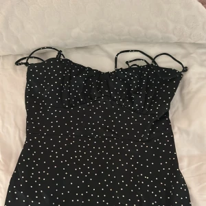 Svart prickig klänning från Zara - Snygg svart klänning från Zara med vita prickar och smala axelband. Klänningen har en elegant slits och är perfekt för en stilren look. Tillverkad i ett lätt och bekvämt material.