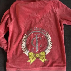 Rosa kofta från Juicy Couture - Säljer en rosa kofta från Juicy Couture med glittrig logga och gul rosett på ryggen. Tröjan har dragkedja och fickor framtill, samt en mysig huva. Perfekt för en avslappnad stil.