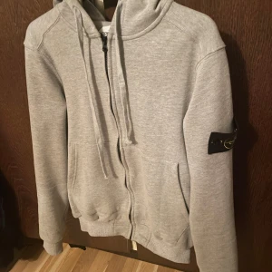 Grå hoodie från Stone Island - Säljer en stilren grå hoodie från Stone Island med dragkedja och huva. Tröjan har en klassisk design med ribbade muddar och en ikonisk logotyp på ärmen. Perfekt för en avslappnad look.