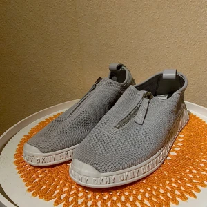 Grå sneakers från DKNY - Snygga grå sneakers från DKNY med dragkedja framtill och stickad ovandel. Skorna har en vit sula med DKNY-logga runtom och på undersidan. Perfekta för en stilren och bekväm look.