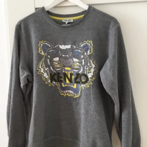 Grå sweatshirt från Kenzo med tigerbroderi - Säljer en grå sweatshirt från Kenzo med ett coolt tigerbroderi i blått, gult och vitt på framsidan. Storleken är M och passar bra till m i passform hade it rekommenderat om du har L som storlek!