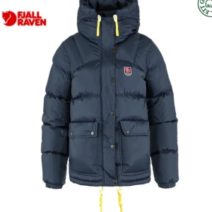 Mörkblå dunjacka från Fjällräven - Säljer en snygg och varm mörkblå dunjacka från Fjällräven. Jackan har en praktisk huva och gula detaljer som ger en cool kontrast. Perfekt för kalla vinterdagar med sina långa ärmar och rejäla fickor. Passar både till vardags och utomhusaktiviteter. 🧥❄️