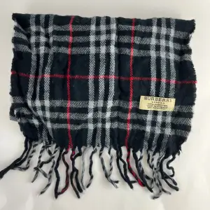 Säljer en snygg rutig halsduk från Burberry i 100% lammull. Den har en klassisk design med svarta, grå och röda rutor. Perfekt för att hålla sig varm under kyliga dagar. Halsduken är i mycket bra skick och har fransar i ändarna. Passar perfekt till både höst och vinteroutfits!