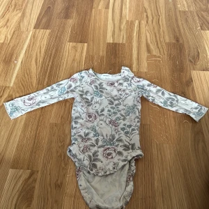Blommig body från Newbie - Söt blommig body från Newbie i storlek 68 för bebisar 4-6 månader. Den har ett vackert blommönster i dova färger och långa ärmar. Perfekt för mysiga dagar.