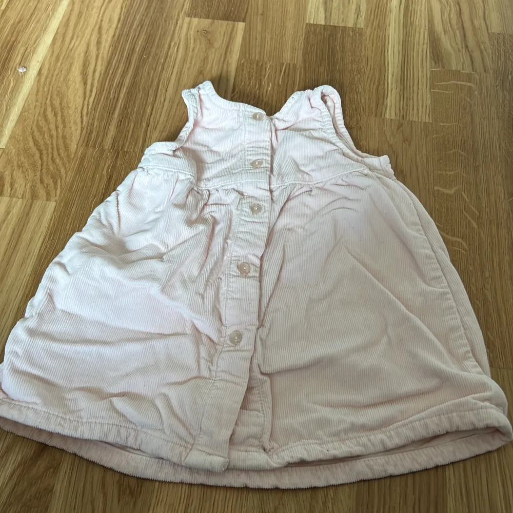 Söt ljusrosa ärmlös klänning för bebis med knappar på baksidan och två fickor framtill. Perfekt för varma dagar och enkel att ta på och av. Tillverkad i mjukt material för extra komfort.. Mekot.