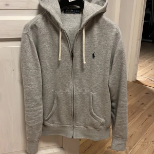 Grå hoodie från Polo Ralph Lauren - Säljer en snygg och bekväm grå hoodie från Polo Ralph Lauren. Den har en dragkedja framtill och en klassisk broderad logga på bröstet. Perfekt för både vardag och chill. Huvan har justerbara snören och tröjan har praktiska fickor. Pris 900kr