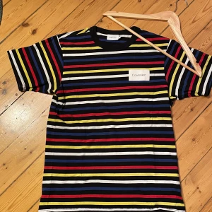 Randig t-shirt från Calvin Klein - Säljer en snygg randig t-shirt från Calvin Klein i storlek M. Den har ett coolt mönster med ränder i svart, vit, blå, röd och gul. Perfekt för en avslappnad stil och passar till både vardag och fest. T-shirten är i mycket bra skick och gjord av mjuk bomull.