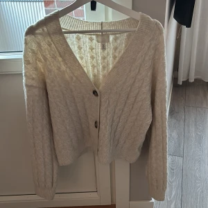 Vit stickad kofta med knappar - Mysig vit stickad kofta med två knappar framtill. Koftan har ett klassiskt flätmönster och långa ärmar