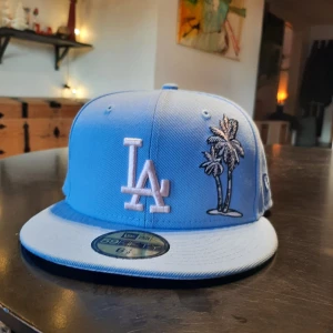 Ljusblå keps från New Era - Snygg ljusblå keps från New Era med LA-logga och palm broderad på framsidan. Kepsen har en rak skärm och är en del av 59FIFTY-serien. Perfekt för en avslappnad stil.