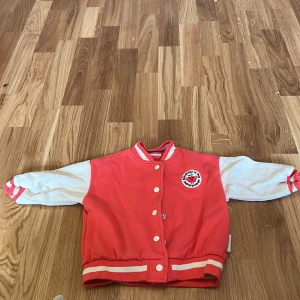 Röd och vit bomberjacka från Minories - Söt bomberjacka för barn från Minories i rött och vitt med broderat märke 'Kindness is my superpower'. Jackan har vita ärmar och knappar framtill samt ribbade muddar i rött och vitt.