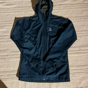 Höglöfs jacka blå - Säljer en mörkblå jacka från Haglöfs i storlek S. Jackan har en praktisk huva och dragkedja framtill. Perfekt för regniga dagar och utomhusaktiviteter.  Nypris: ca 1400. Mitt pris:499
