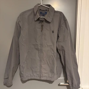Grå jacka från Polo Ralph Lauren - Säljer en stilren grå jacka från Polo Ralph Lauren. Den har en dragkedja framtill och en klassisk krage. Jackan är i bra skick och passar perfekt för höst och vår. Den har långa ärmar och elastiska muddar vid ärmslut och nederkant. Perfekt för en casual look. Den är sprilands ny köpt för 2700