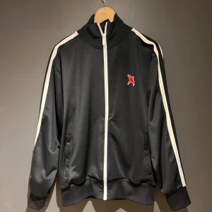 Svart träningsjacka från Axel Arigato - Snygg svart träningsjacka från Axel Arigato med vita ränder längs ärmarna och en röd logga på bröstet. Jackan har dragkedja framtill och är tillverkad i polyester med lite elastan för extra komfort.