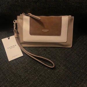 Väska / plånbok  - ULRIKA DESIGNKLASSISK COLOR PALETTE PURSE/WALLET.                                                                         Snygg och praktisk plånbok från Ulrika i beige och brun färg. Den har en dragkedja och flera fack för kort. Perfekt för att hålla ordning på dina saker. Nytt skick med etikett kvar. Perfekt för både vardag och fest!           Mått: H 19cm x B 10,5cm.  Nypris: 399:-