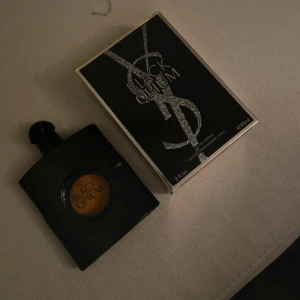 Black Opium Eau de Parfum från Yves Saint Laurent - Säljer en flaska Black Opium Eau de Parfum från Yves Saint Laurent. Flaskan är svart med glittrande yta och har en rund öppning som visar parfymens gyllene färg. Förpackningen är elegant med YSL-loggan i silver och texten 'Black Opium'. Perfekt för den som älskar en djärv och sensuell doft.