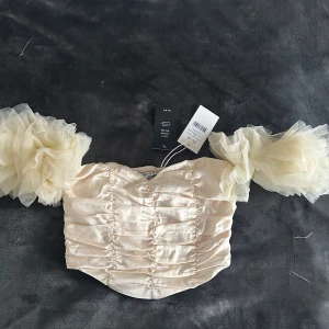 Krämfärgad topp med volangärmar från NA-KD - Säljer en elegant krämfärgad topp från NA-KD med volangärmar i tyll. Toppen har en rynkad design framtill och är perfekt för en stilfull look. Den är axelbandslös och har en kort passform. Oanvänd💞