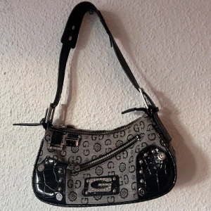 GUESS baguette bag - Snygg axelväska med ett unikt mönster i svart och grått. Väskan har svarta detaljer i skinnimitation och silverfärgade nitar. Den har en dragkedja upptill och justerbar axelrem. Märke Guess men inte helt säker på om den är äkta.