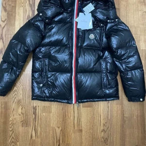 Svart dunjacka från Moncler - Snygg svart dunjacka från Moncler med en glansig finish och ikonisk logga på bröstet. Jackan har en praktisk huva och stängs med en dragkedja i rött, vitt och blått. Perfekt för kyliga dagar.