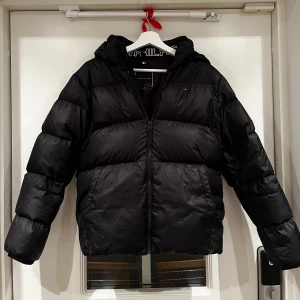 Svart pufferjacka från Tommy Hilfiger - Snygg svart pufferjacka från Tommy Hilfiger med huva och dragkedja. Perfekt för kalla dagar med sin vadderade design. Jackan har en klassisk look och är långärmad.