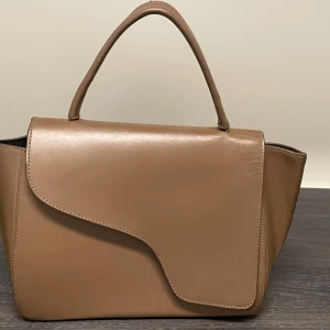 ATP Atelier Arezzo handväska Almond - Arezzo handväska är handgjord av vegetabiliskt garvat Vacchetta-läder. Väskan är i färgen ”almond” och har en avtagbar axelrem. I använt men välvårdat skick. Den har några smårepor som knappt syns, se bild 5. Kommer med dustbag. Storlek Höjd 18,m cm, Bredd 34 cm, Djup 10,5 cm. 