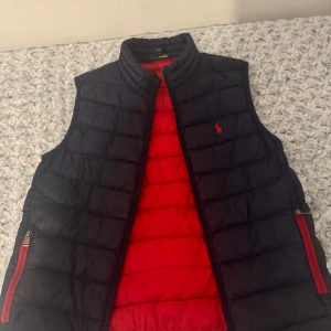 Svart och röd dunväst från Polo Ralph Lauren - Säljer en snygg svart dunväst från Polo Ralph Lauren med röd insida. Västen har en klassisk design med dragkedja och är perfekt för höst och vår. Den är i mycket bra skick och passar både till vardags och lite finare tillfällen.