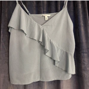 Grå ärmlös topp från H&M - Snygg grå ärmlös topp från H&M med volangdetaljer. Perfekt för sommardagar eller en avslappnad kväll. Toppen har en omlott-design och tunna axelband.