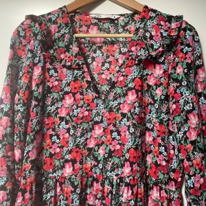 Blommig långklänning från Zara - Superfin blommig långklänning från Zara i storlek S. Klänningen har ett färgglatt mönster med rosa och röda blommor på en svart bakgrund. Den har långa ärmar och en bekväm passform, perfekt året runt. Materialet känns lätt och luftigt, vilket gör den idealisk för alla kroppsformer. Inga defekter på varan. Som ny.