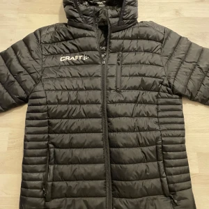 Svart pufferjacka från Craft - Säljer en svart lättvikt/pufferjacka från Craft. Nypris 999kr. Den är perfekt för kyliga dagar med sin quiltade design och långa ärmar. Jackan har en dragkedja framtill och en praktisk huva. Den är i bra skick och redo för nya äventyr!