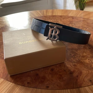 Svart bälte från Burberry - Säljer ett snyggt svart bälte från Burberry med deras stilrena mönster. Bältet har ett glänsande silverfärgad spänne med Burberrys ikoniska logga. Perfekt för att ge en lyxig touch till vilken outfit som helst. Kommer i originalförpackning. 100cm