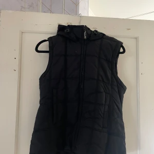 Svart dunväst från H&M - Säljer en svart dunväst från H&M i storlek XS. Västen har en dragkedja framtill och en praktisk huva. Perfekt för kyliga höst- och vårdagar. Den är quiltad och har en normal passform.