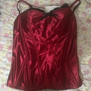 Röd korsett från Body Zone - Snygg röd korsett från Body Zone med svarta spetsdetaljer och en liten rosett framtill. Den har justerbara axelband och en glansig finish som ger en lyxig känsla. Lite stor på mig som brukar ha 75b, men den passar fortfarande bara inte lika ”snatched” men går lätt att sy in för dne händiga 💗