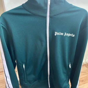 Grön träningsjacka från Palm Angels - Snygg grön träningsjacka från Palm Angels med vit dragkedja och vita ränder längs ärmarna. Jackan har en hög krage och två fickor med dragkedja.